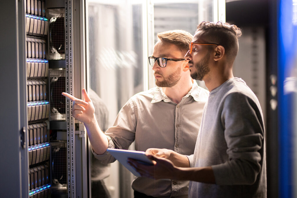 Zwei Netzwerkspezialisten stehen vor Serverschrank im Data Center und konfigurieren über ein Tablet in der Hand das Netzwerk